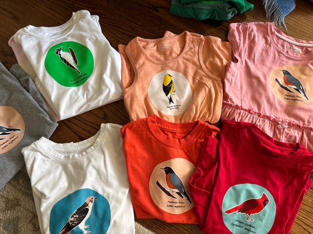 A collection of colorful t-shirts and tank tops featuring bird illustrations and names, arranged on a wooden surface. The shirts include a white t-shirt with a woodpecker graphic, an orange tank top with a goldfinch, and a red t-shirt with a cardinal design.